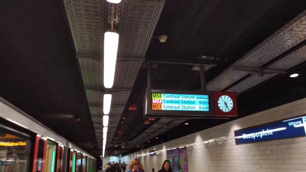 Platform Spoor 2 at metro station Weesperplein with people exiting. Digital signage with an analog clock, showing departures: M54 Centraal Station, M53 Centraal Station 4 min, M51 Centraal Station 8 min.